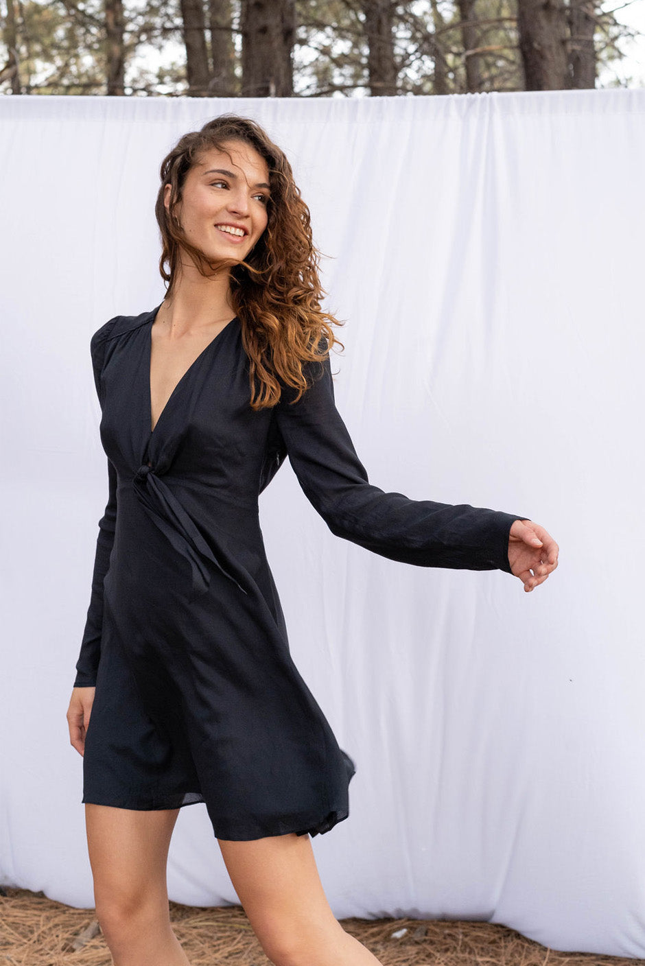 Holly mini dress in noir black color for women by Paneros Clothing. Handmade from sustainable deadstock rayon fabric, with a tie neck front with full sleeves and smocking. Side view.