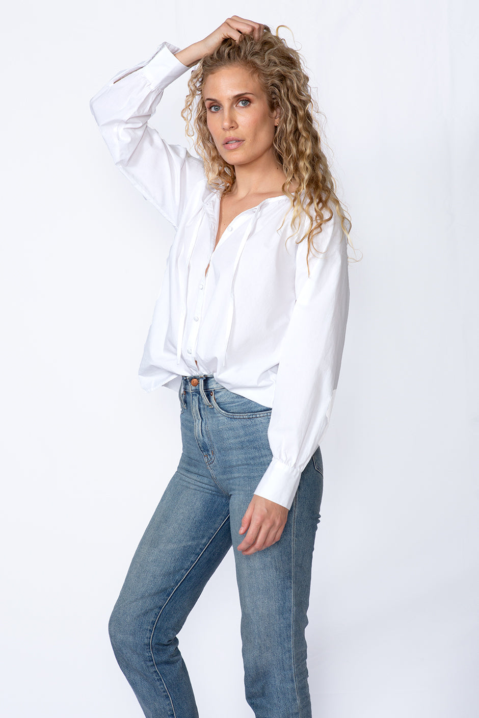 Mesa blouse in cloud white color for women by Paneros Clothing. Handmade from sustainable deadstock cotton poplin fabric, button-up with full sleeves, tie neck, and slight shirttail. Front side view styled with jeans.